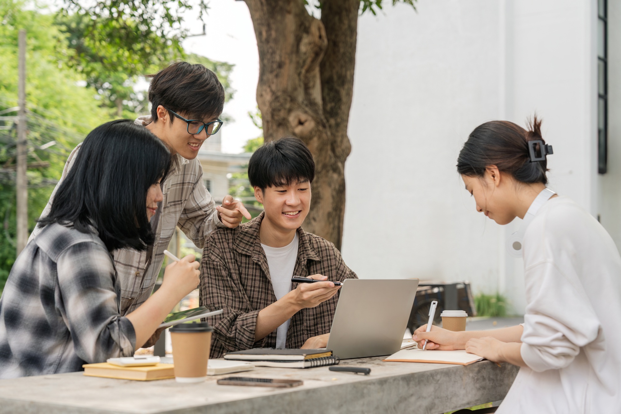 Group of happy young Asian college students sitting on a on the cement table, looking at a laptop